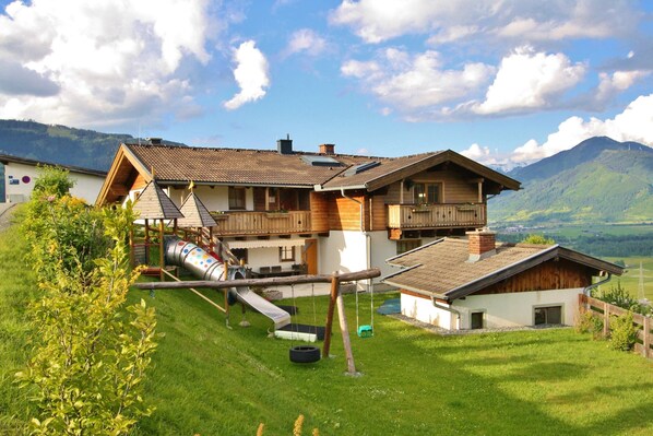 A chalet-style house with a sloped roof, a wooden deck, and a playground area including a slide and swings. - Modern Mansion in Kaprun Near Ski Area (Kaprun)