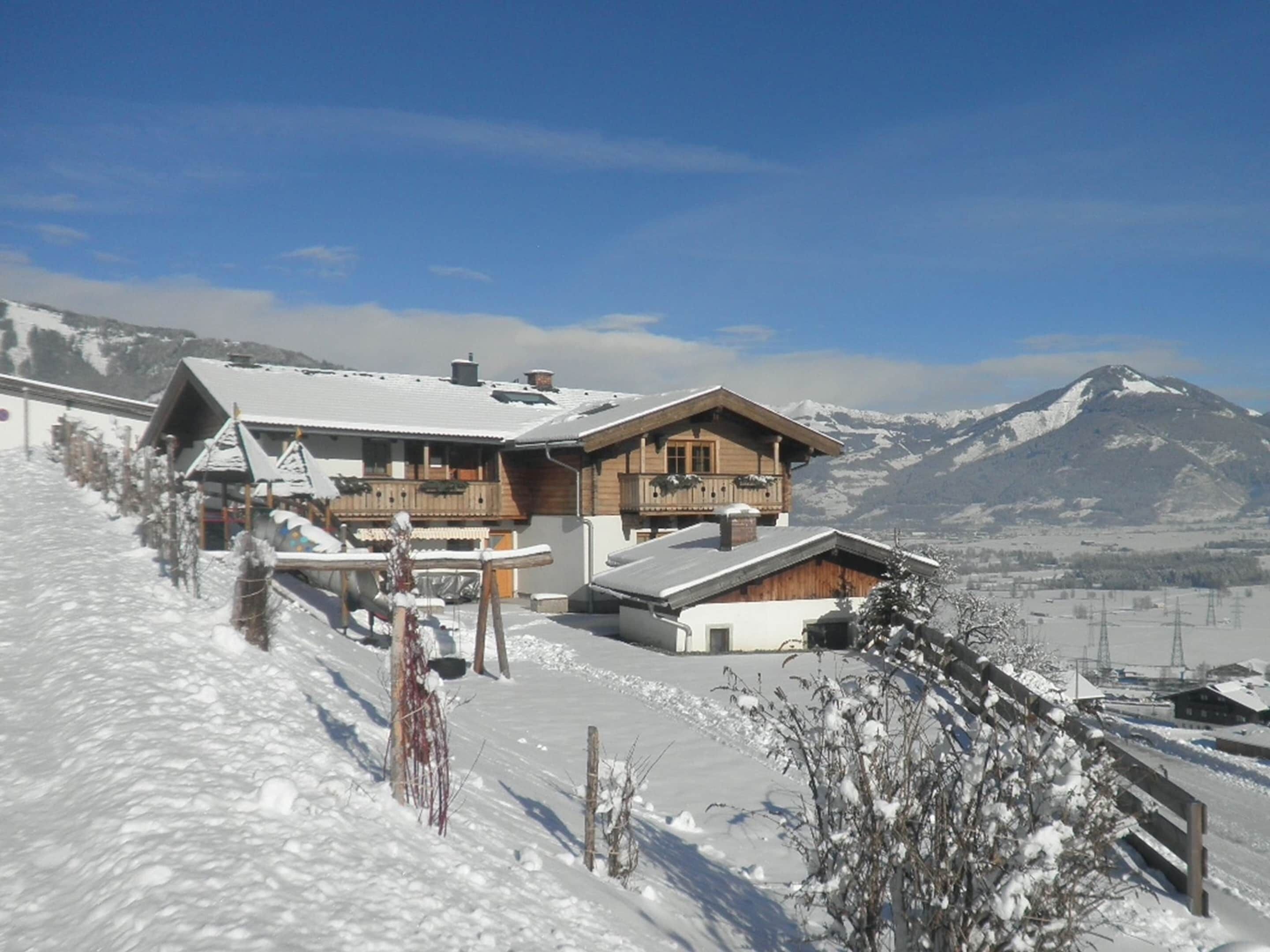 A wooden chalet with a balcony, surrounded by snow-covered landscape and mountains in the background.