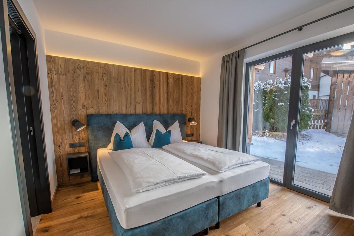 Une chambre moderne avec un mur recouvert de panneaux en bois, un grand lit avec une literie blanche et bleue, et une vue sur un espace extérieur enneigé, visible à travers une porte vitrée.