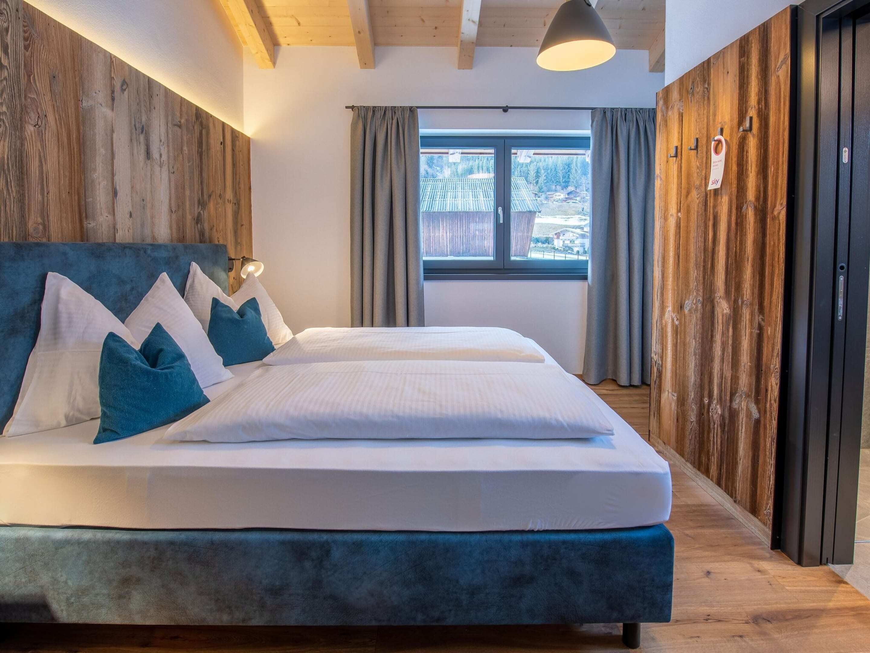 A double bed with white and blue bedding, a wooden headboard, and a large window with curtains.