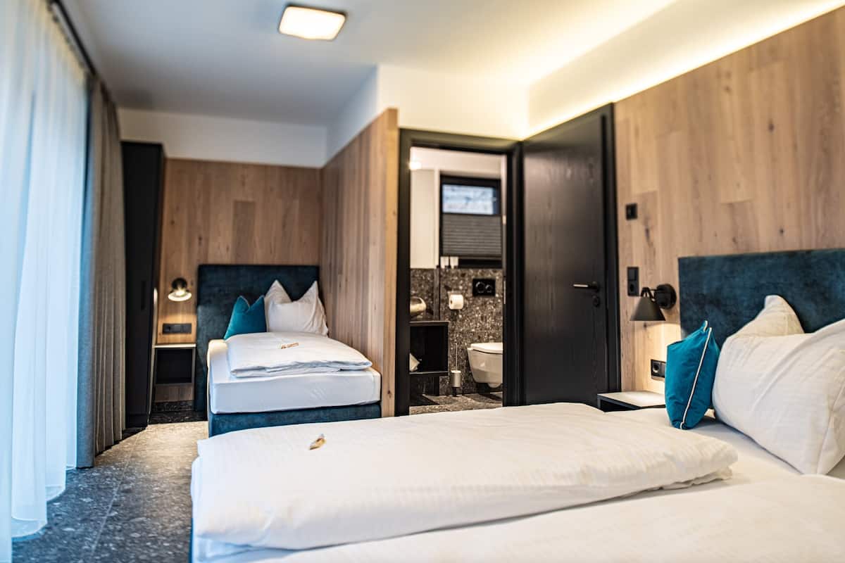 Une chambre d’hôtel avec deux lits, une tête de lit en bois, une petite table de chevet et une commode, ainsi qu’une salle de bain visible à travers une porte ouverte.