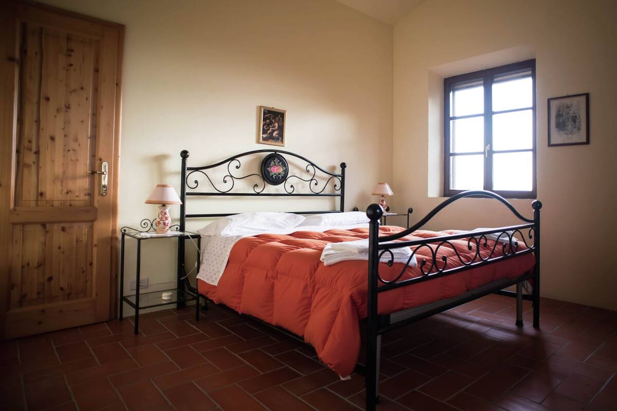 A bedroom with a metal bed frame, a wooden door, a window, and a small bedside table with a lamp.