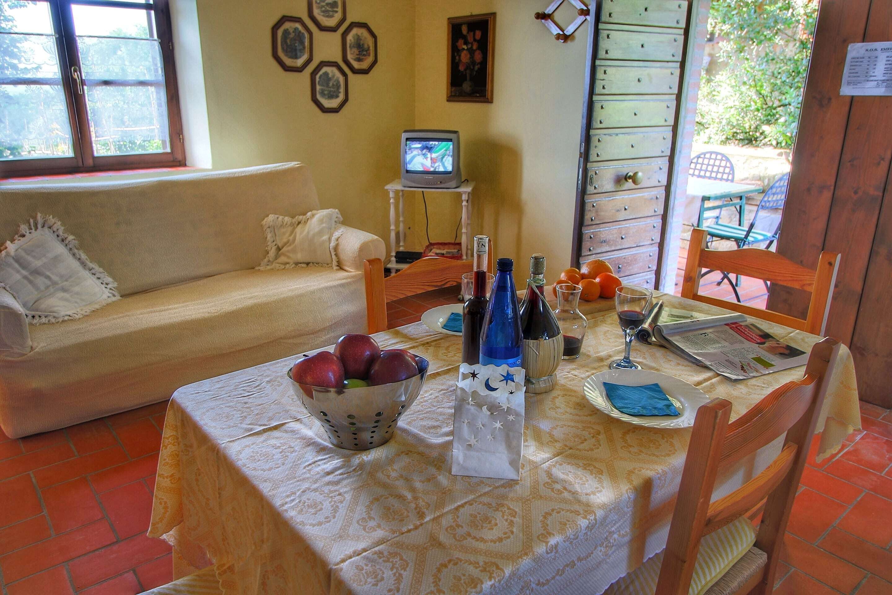 A dining area with a table set for two, a television on a stand, and a window with a view of greenery.