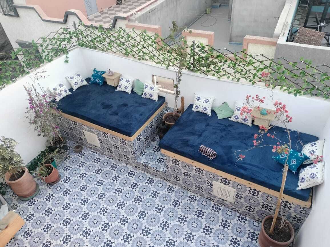A rooftop terrace with two blue cushioned seating areas, potted plants, and a patterned floor.