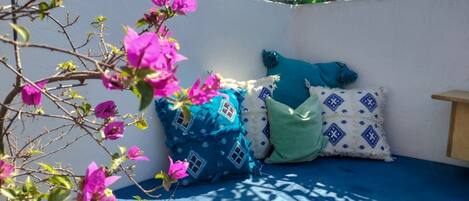 A room with a blue bed, patterned pillows, and a plant with pink flowers.