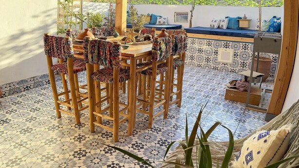 Un patio techado con una mesa y sillas de madera, piso de baldosas y vistas a un jardín y edificios al fondo.