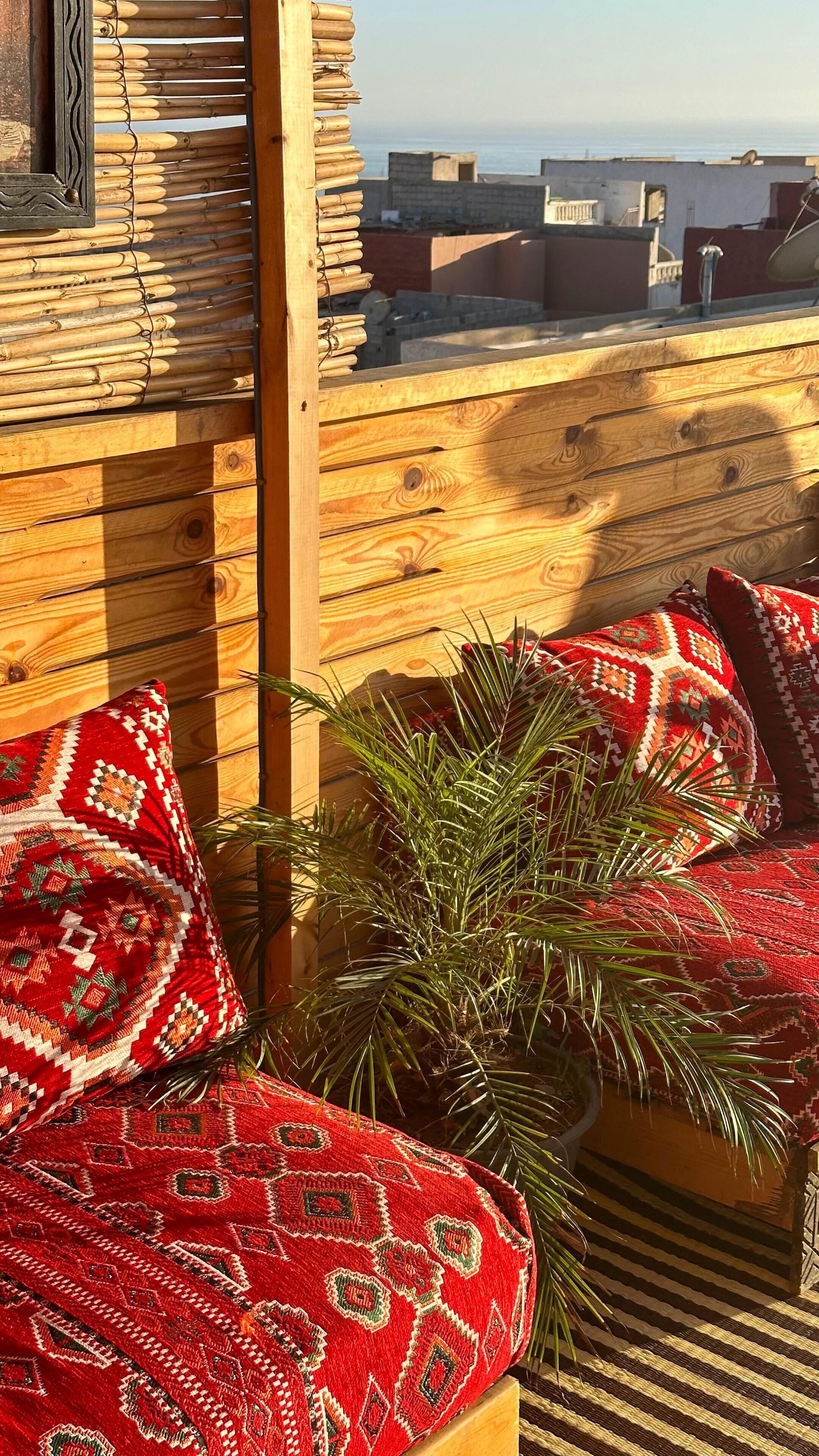 A wooden balcony with a view of the sea, a palm plant, and red patterned cushions.
