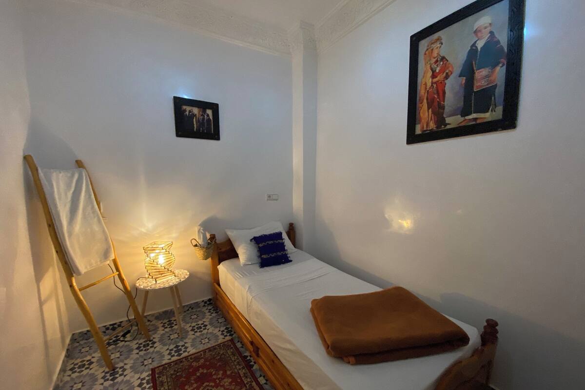 Une petite chambre à coucher, bien rangée, avec un lit simple, une échelle en bois, une petite table avec une lampe et un tableau encadré au mur.