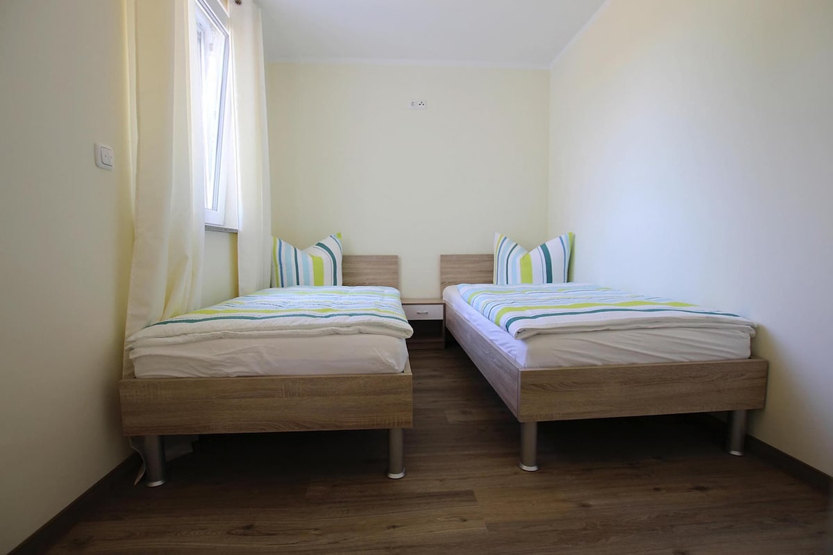 A small, clean room with two wooden beds, a window with curtains, and a light switch on the wall.