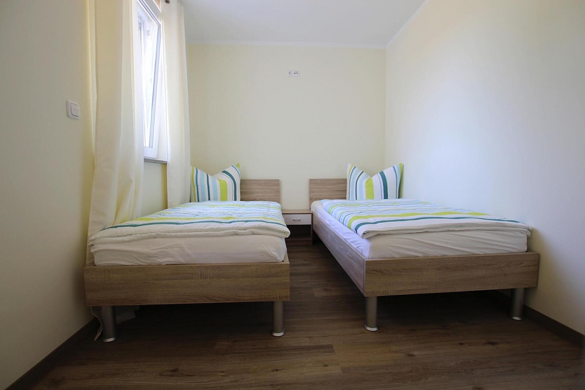 A small, clean room with two wooden beds, a window with curtains, and a light switch on the wall.