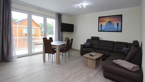 Un salon moderne avec un canapé marron, une table basse en bois et une table à manger avec des chaises. Un téléviseur à écran plat est fixé au mur, et il y a une grande fenêtre avec des rideaux.