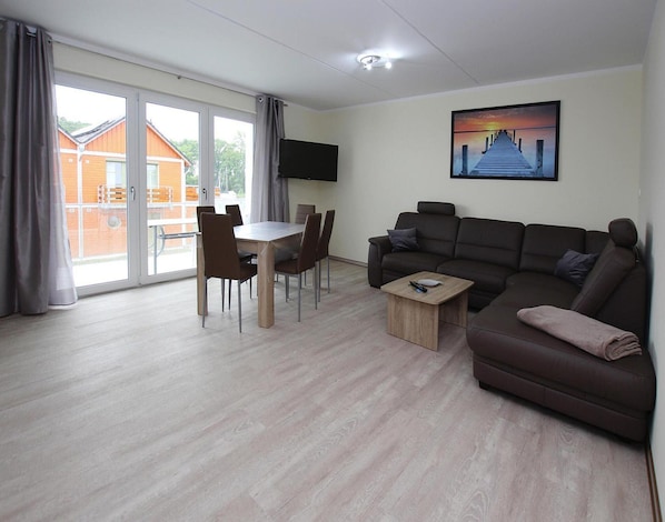 A modern living room with a brown sofa, a wooden coffee table, and a dining table with chairs. A flat-screen TV is mounted on the wall, and there is a large window with curtains. - Apartment Hafenflair am Plauer See, Plau am See (Plau am See)