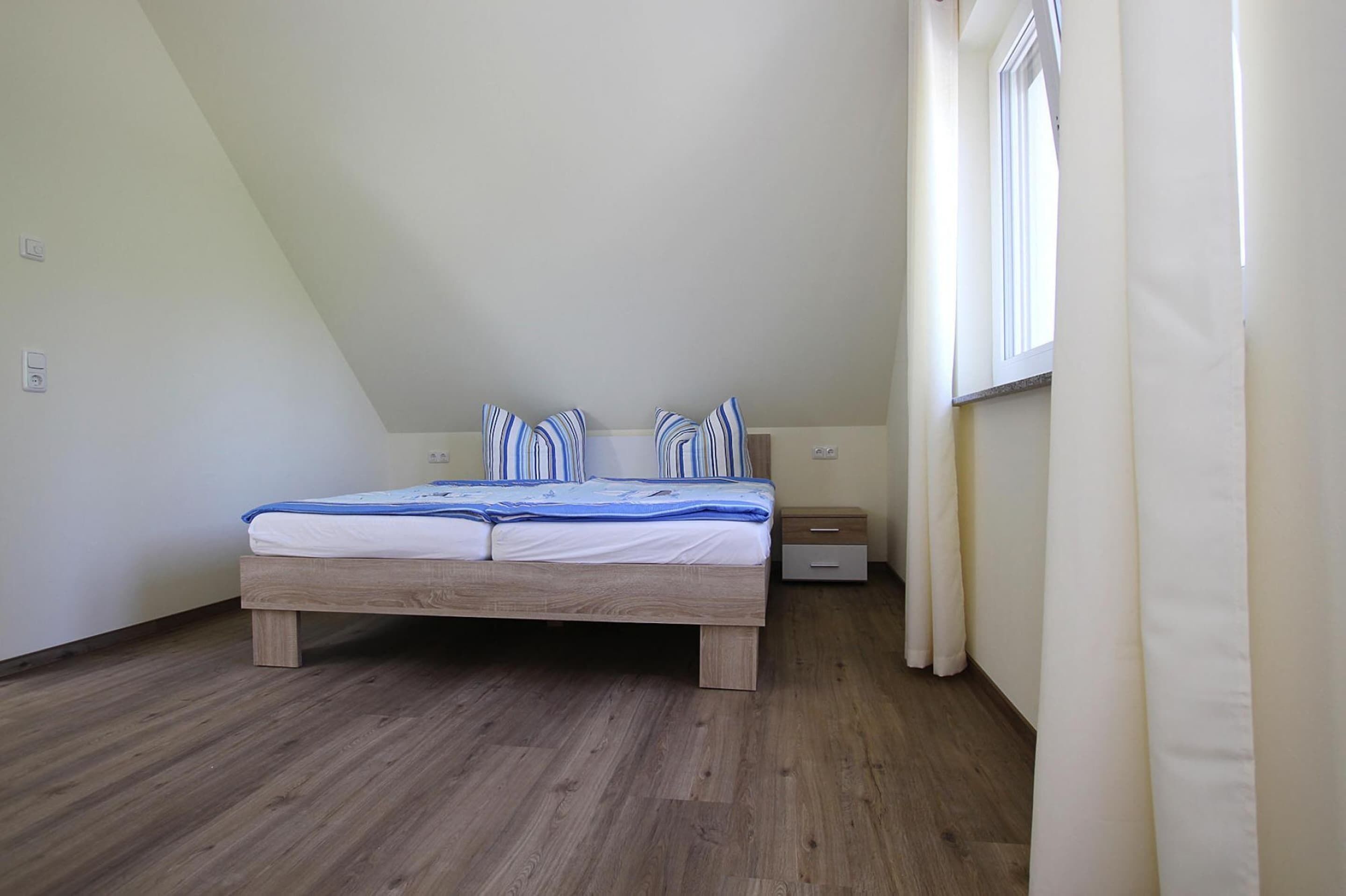 A bedroom with a wooden bed, a nightstand, a window with curtains, and a sloped ceiling.