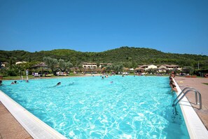 A large swimming pool with clear blue water, surrounded by lounge chairs and umbrellas, with buildings and greenery in the background.