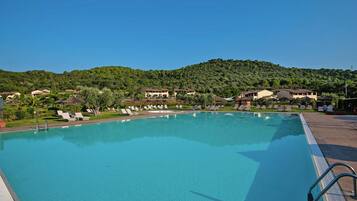 A large, clear swimming pool with lounge chairs and a view of a hillside with buildings.