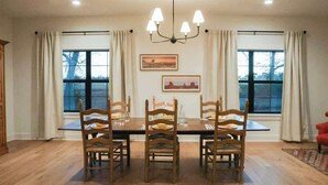 A dining room with a large wooden table, six chairs, a chandelier, two windows with curtains, and framed pictures on the wall. - Rest & Relax | Pool | Countryside View | Sleeps 6 (Fayetteville)