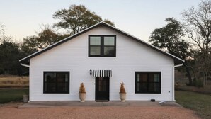 A white single-story house with a gabled roof, a front door, and two large windows on either side. - Rest & Relax | Pool | Countryside View | Sleeps 6 (Fayetteville)