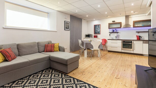 Una sala moderna con un sofá seccional gris, una zona de comedor con mesa y sillas, una cocina con armarios blancos y un horno de acero inoxidable, y una alfombra estampada en el suelo.