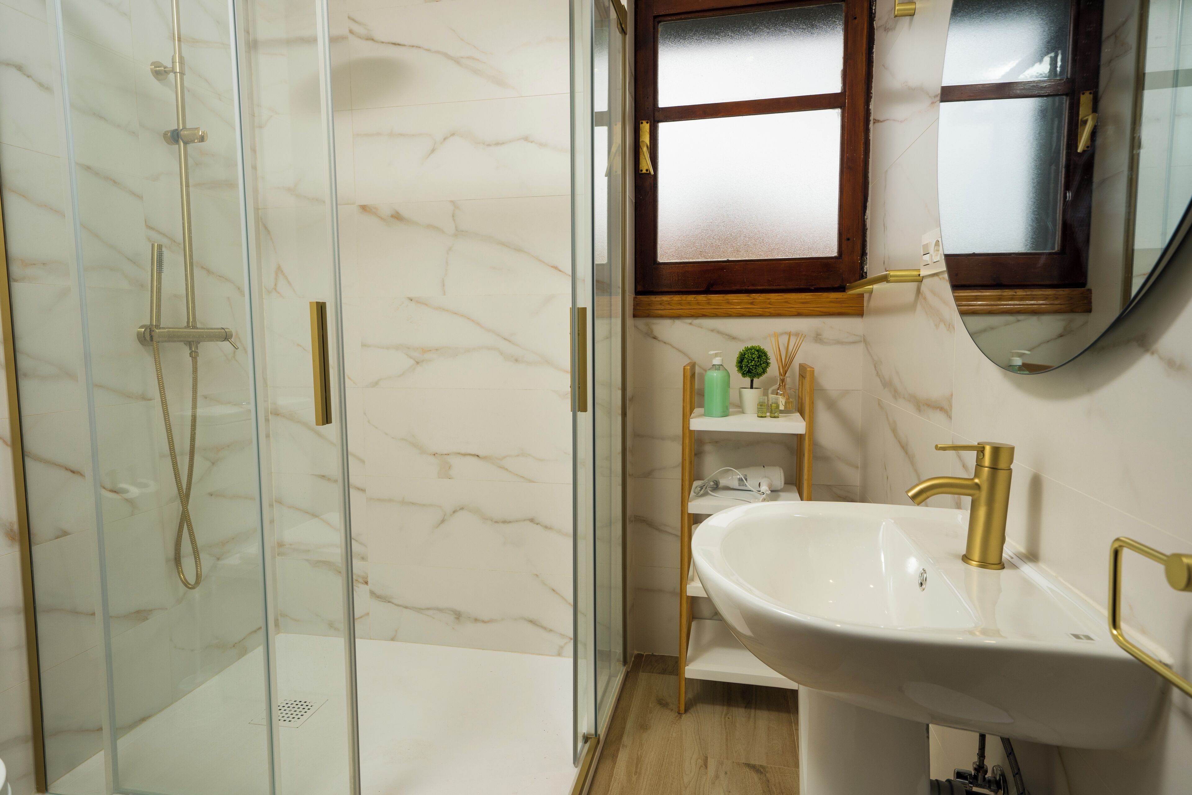A bathroom with a glass-enclosed shower, a white sink with gold fixtures, and a wooden vanity.