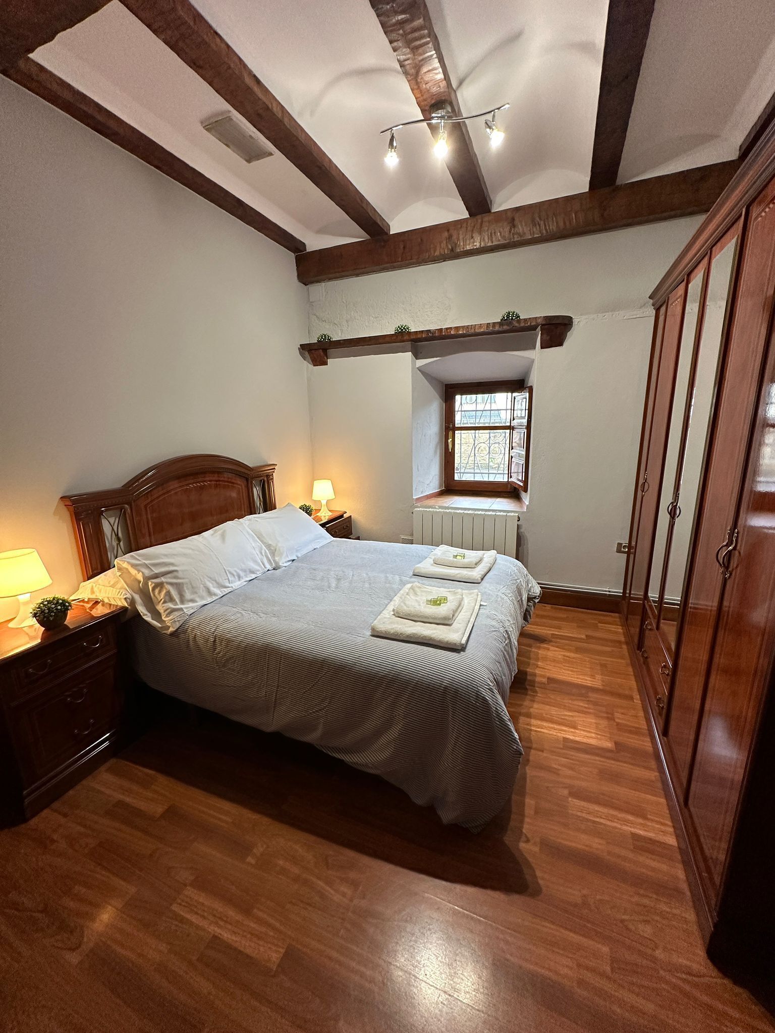 A bedroom with a wooden bed, a nightstand with a lamp, a wardrobe, and a window with a view.