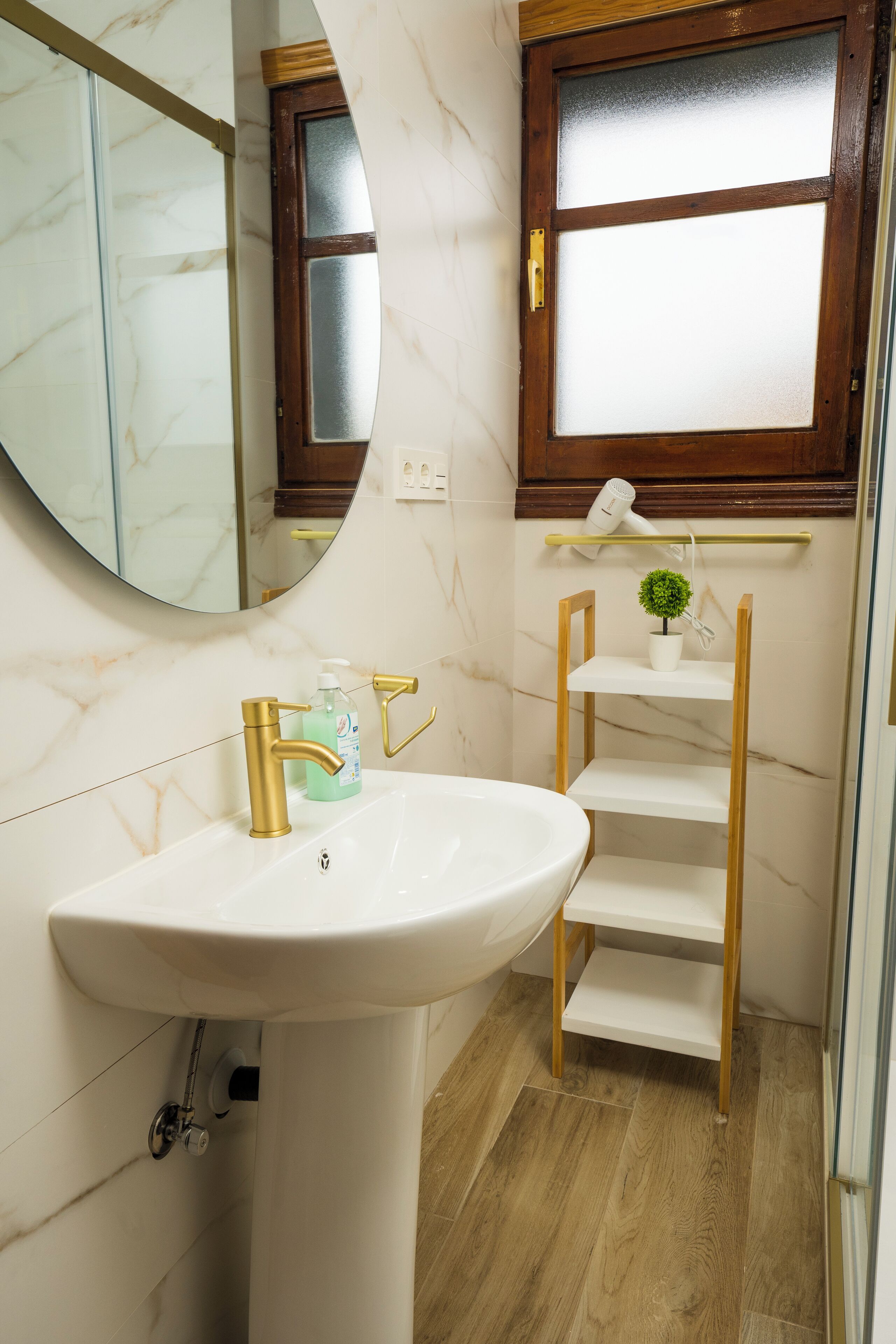 Ein Badezimmer mit einem weißen Waschbecken, goldenen Wasserhähnen, einem Spiegel, einer Holzablage mit einer Pflanze und einem Milchglasfenster.