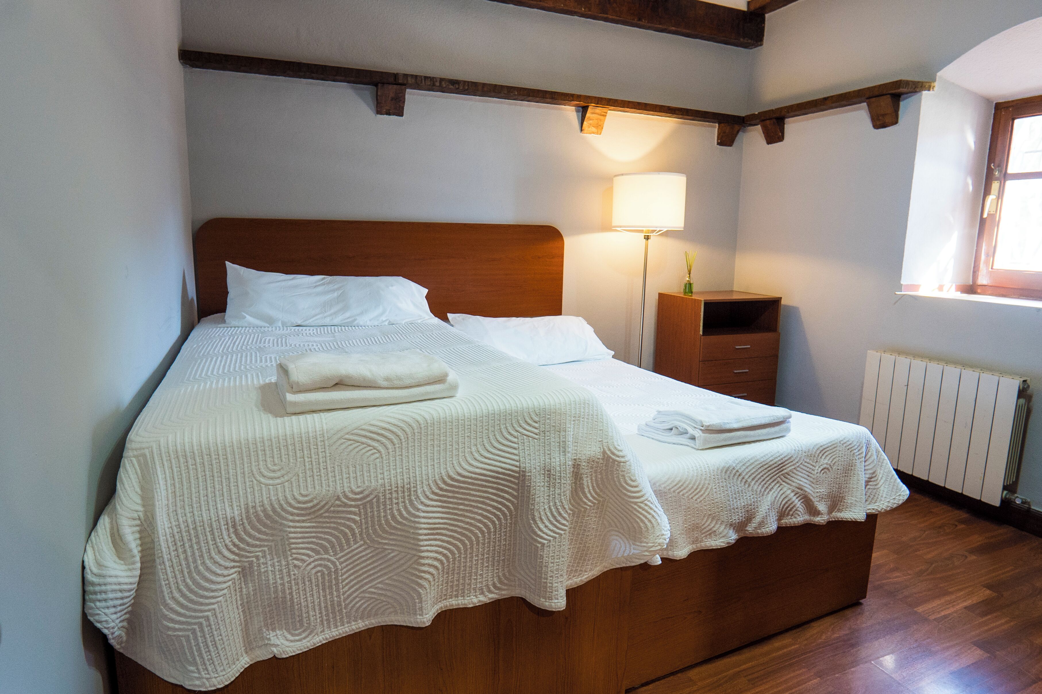 Una cama bien hecha con una manta blanca y almohadas, una lámpara de noche y una mesita de noche de madera con un cajón.