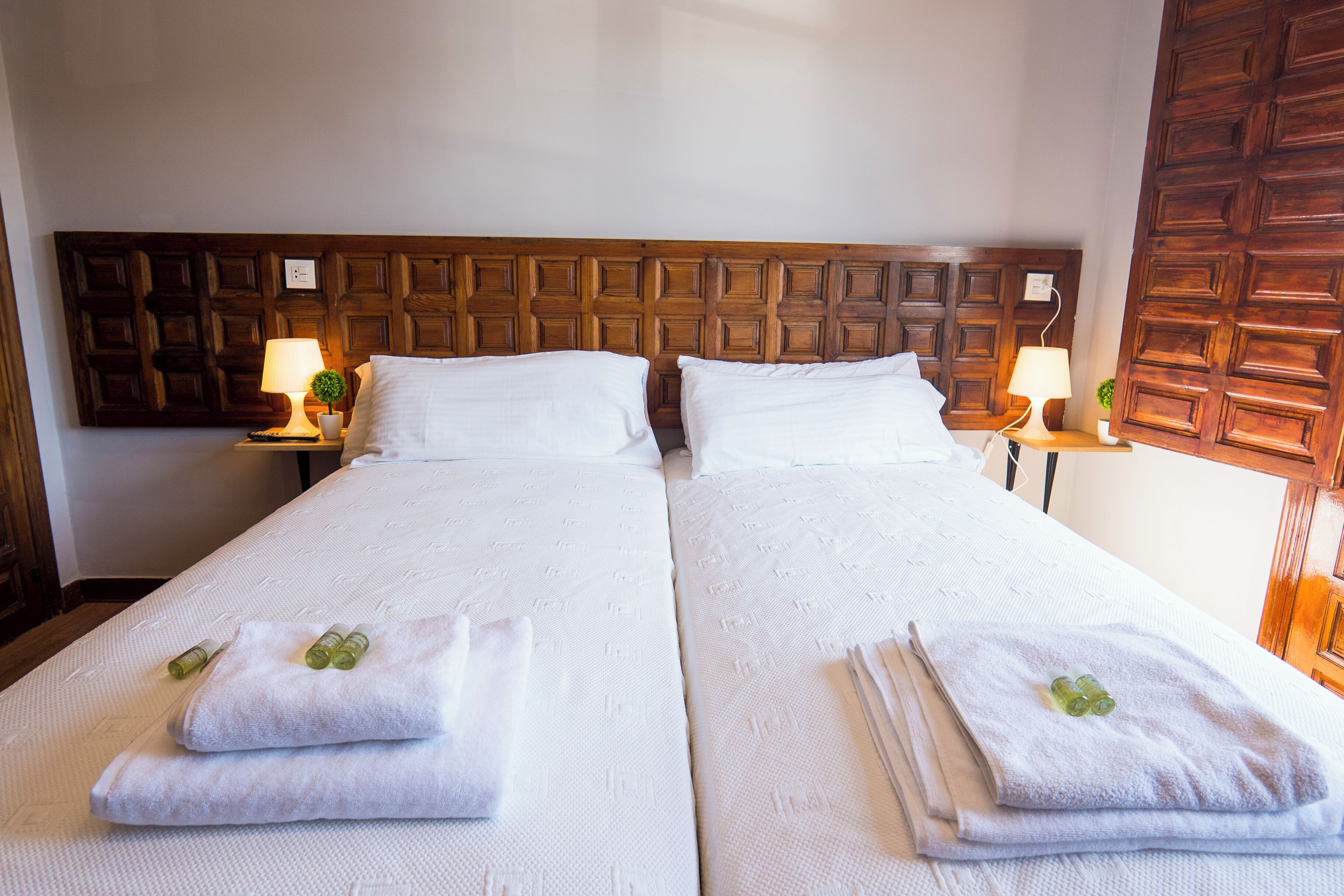Una cama doble con sábanas blancas y dos almohadas, un cabecero de madera, dos mesitas de noche con lámparas y una puerta de madera.