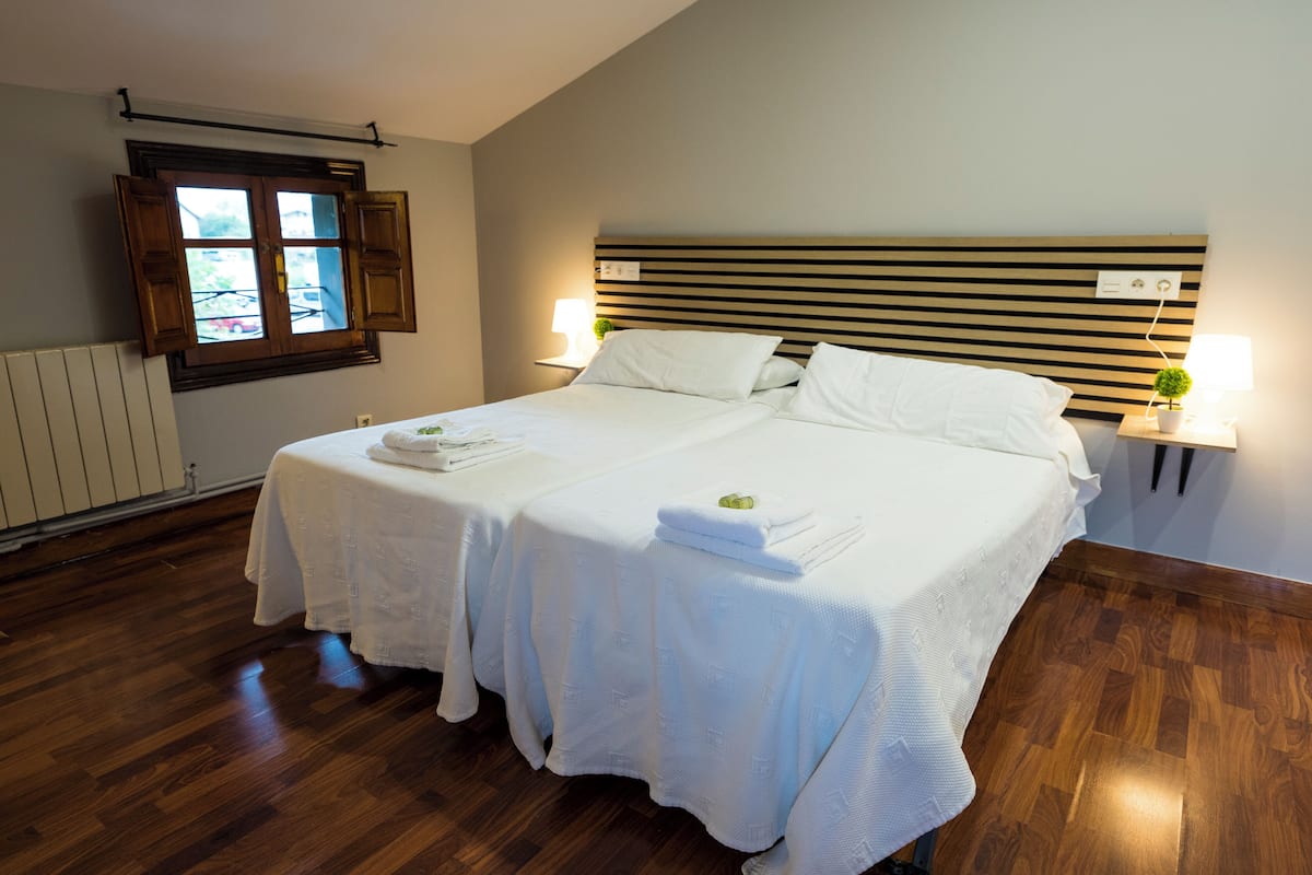 Quarto de hotel com uma cama grande, duas mesas de cabeceira, uma janela com persianas de madeira e um radiador na parede.