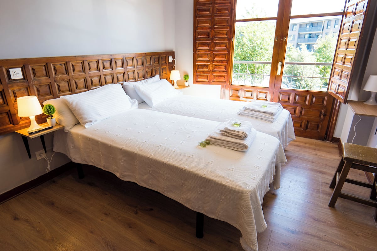 Quarto de hotel com duas camas, cabeceira de madeira, janela com vista para edifícios, uma pequena mesa com luminária e uma cadeira.