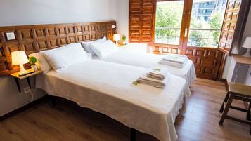Habitación de hotel con dos camas, cabecero de madera, ventana con vistas a edificios, una mesita con una lámpara y una silla.