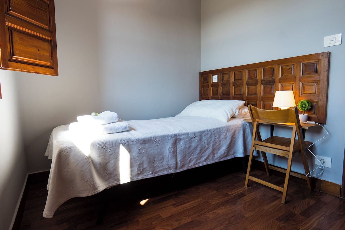 Uma cama de solteiro com cabeceira de madeira, mesa de cabeceira com abajur e uma pequena planta.