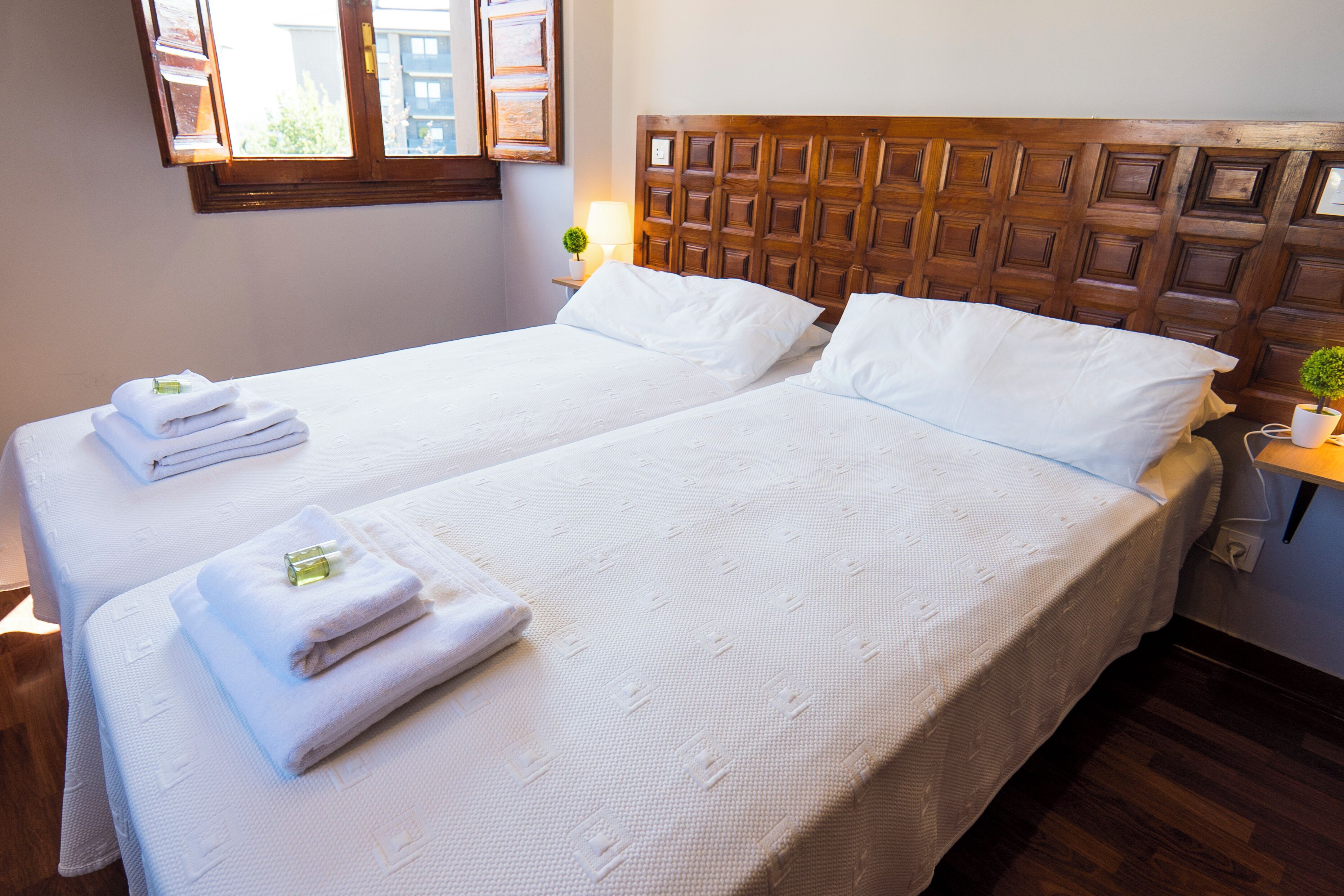 A double bed with white linens and towels, a wooden headboard, a bedside table with a lamp, and a window with blinds.