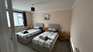 A bedroom with two beds, each with a headboard, a bedside table, and a lamp. There is a window with curtains, a wall-mounted picture, and a radiator. - 2bedroom-1bathroom-patio-wifi (Auchterarder)
