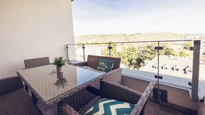 Een balkon met rieten meubels, een glazen tafel en uitzicht op gebouwen en bomen.