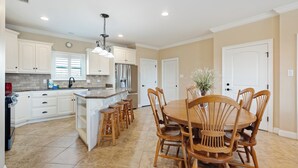 A spacious kitchen with white cabinets, a central island, wooden chairs, and a stainless steel refrigerator.