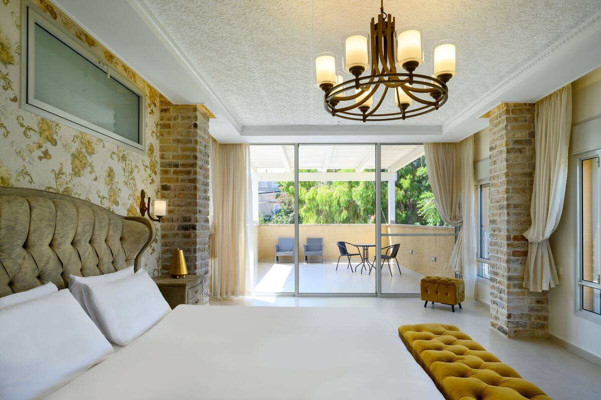 Une chambre spacieuse avec un grand lit, un lustre et un balcon équipé de chaises.
