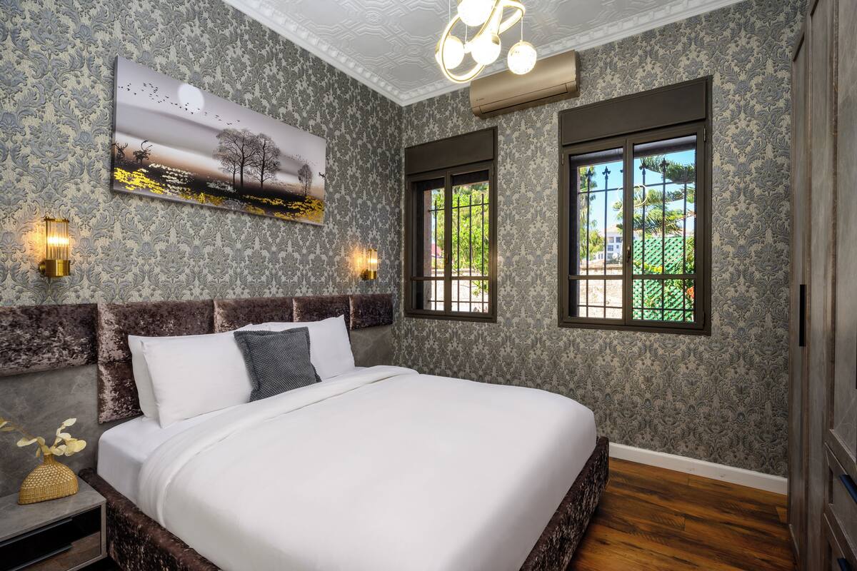 Une chambre à coucher avec un grand lit, un papier peint à motifs, un tableau, deux fenêtres et un luminaire au plafond.