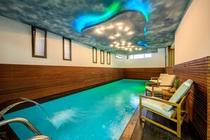 Una piscina interior con paneles de madera, techo con diseño de nubes y algunas sillas de descanso.