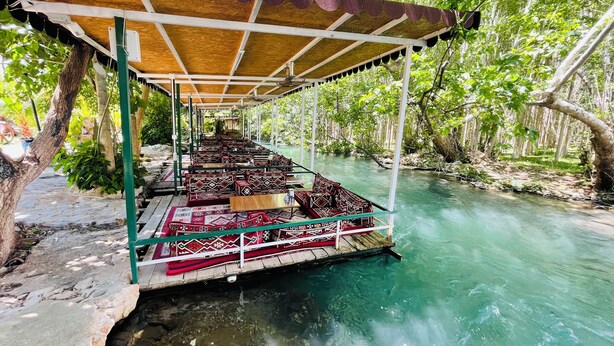A covered seating area with benches and tables by a clear river, surrounded by trees. - biçen hotel (Midyat)