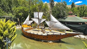 A wooden boat with a covered deck and a small cabin, docked in a calm body of water surrounded by greenery and a clear sky. - biçen hotel (Midyat)
