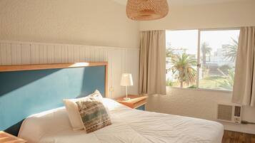 A bedroom with a bed, a bedside table with a lamp, a window with curtains, and a view of palm trees and buildings.