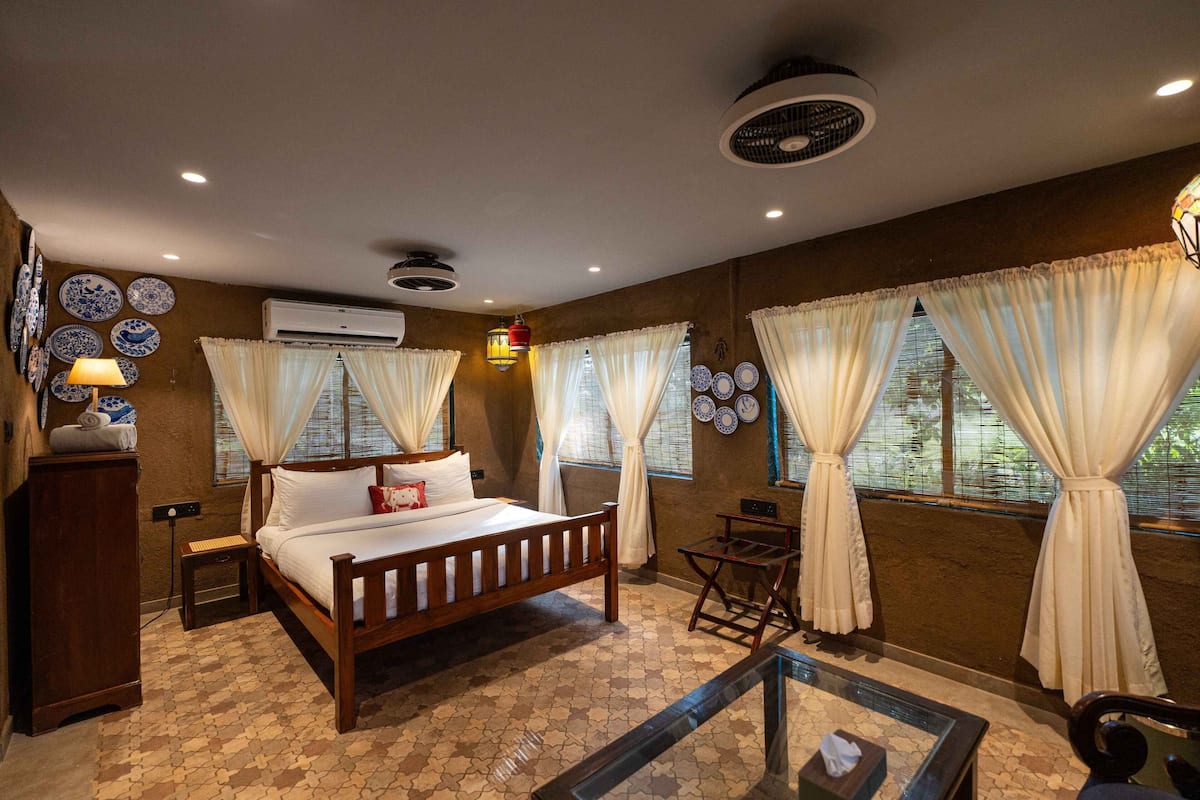 A bedroom with a wooden bed, a small table, and a glass coffee table. There are curtains on the windows and a wall-mounted air conditioner.
