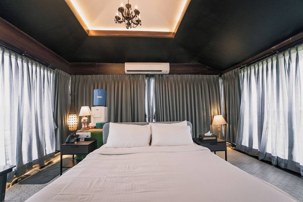 A hotel room with a large bed, two bedside tables with lamps, an air conditioning unit, and a chandelier.