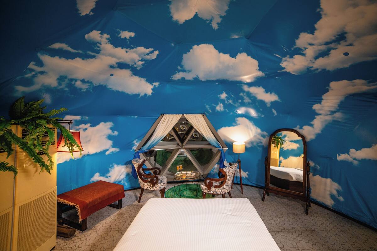 A room with a bed, a small table, two chairs, a bench, a mirror, and a tent-like structure with a lamp.