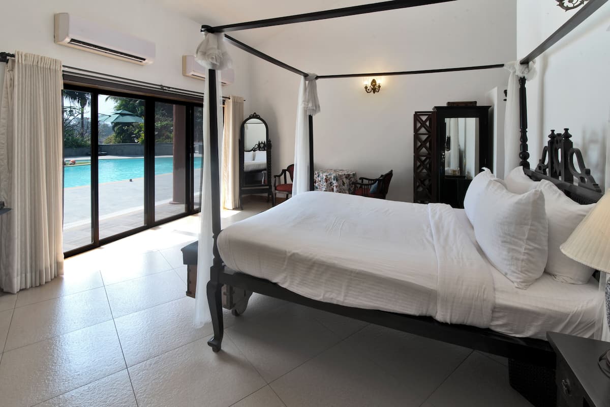 A four-poster bed with white linens, a nightstand with a lamp, a chair, a table with a floral tablecloth, and a large mirror.