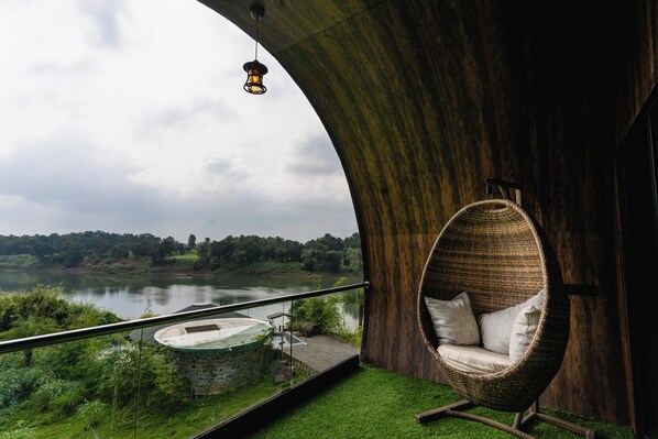 Une chaise en osier avec des coussins, placée sur un balcon donnant sur un lac, et une suspension lumineuse au-dessus.