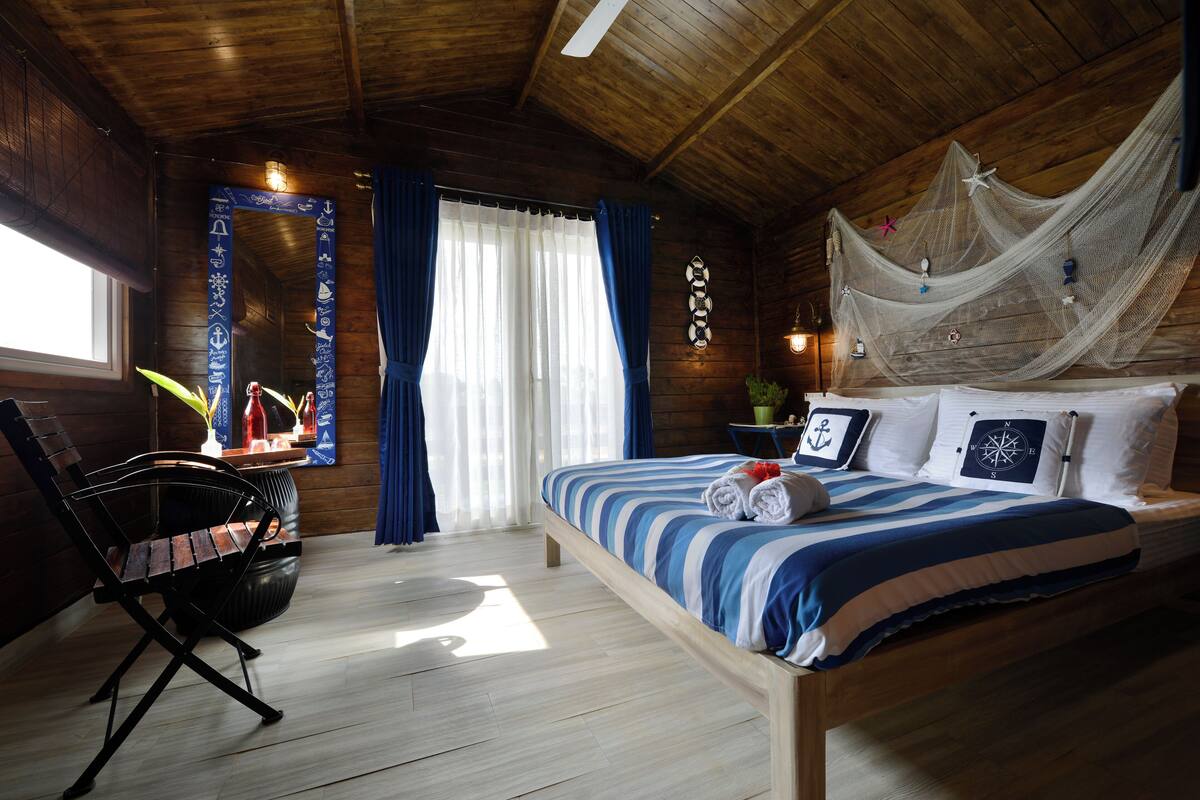 A wooden cabin-style bedroom with a bed, a chair, a window with curtains, and a decorative wall hanging.