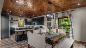 A modern kitchen with wooden ceiling, bar stools, a dining table with benches, and a large window with curtains.