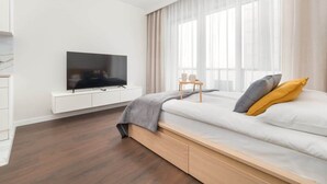 Ein modernes Schlafzimmer mit einem Bett, einem Fernseher auf einer weißen Kommode und einem Fenster mit leicht transparenten Vorhängen.