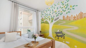 A bedroom with a bed, a bedside table with a vase of flowers, a window with curtains, and a wall mural of a tree and landscape. - Spacious Apartment for 6 Persons | Parking | Workspace (Kraków)